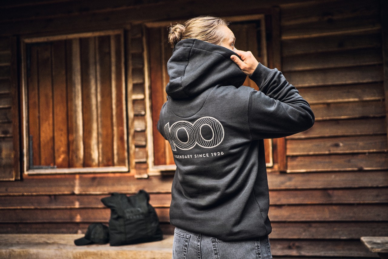 Mujer con la sudadera "STIHL 100" de la colección aniversario frente a una cabaña de madera.