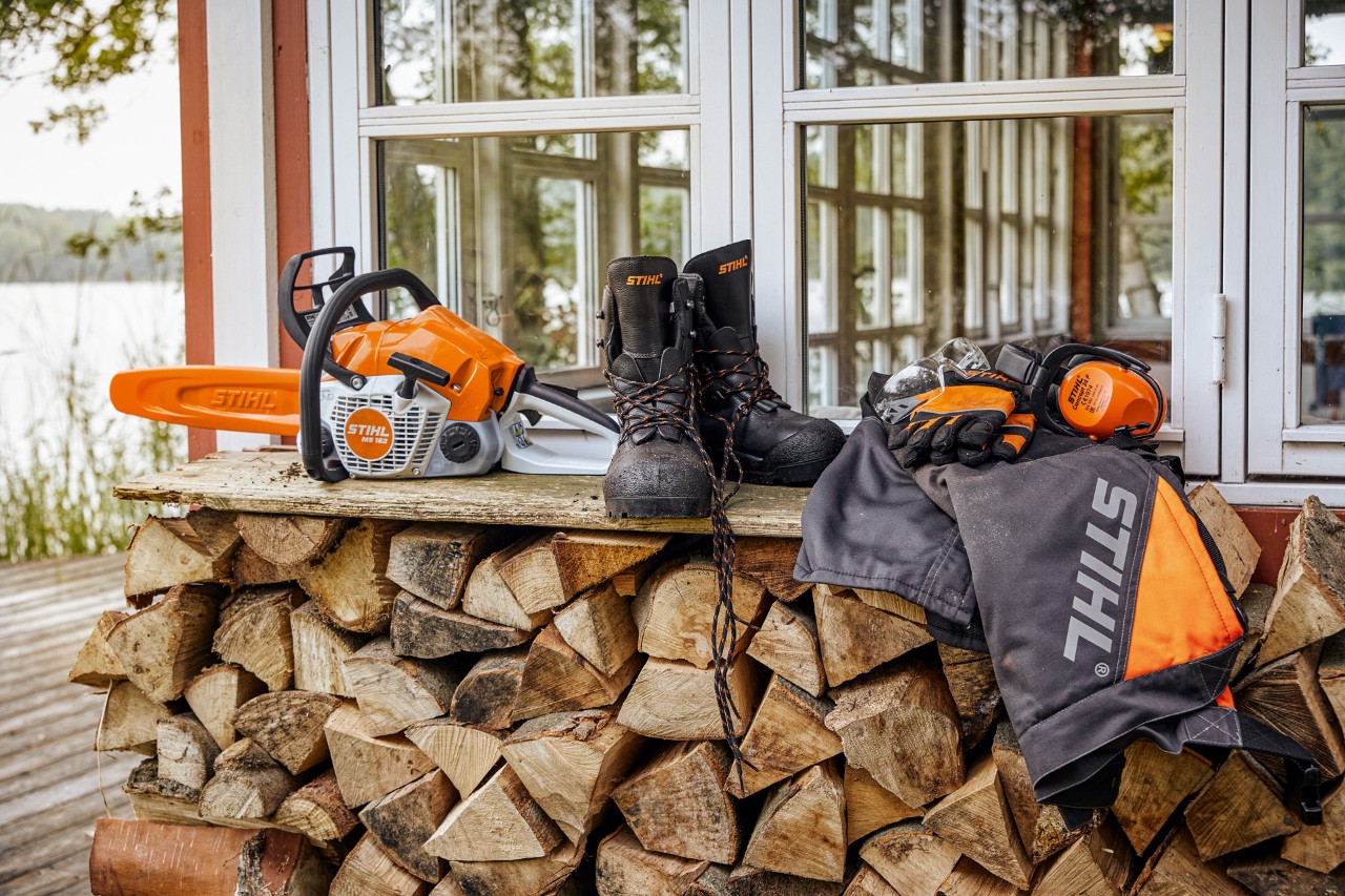 En una estantería sobre leña apilada, hay ropa de protección STIHL y la MS 162