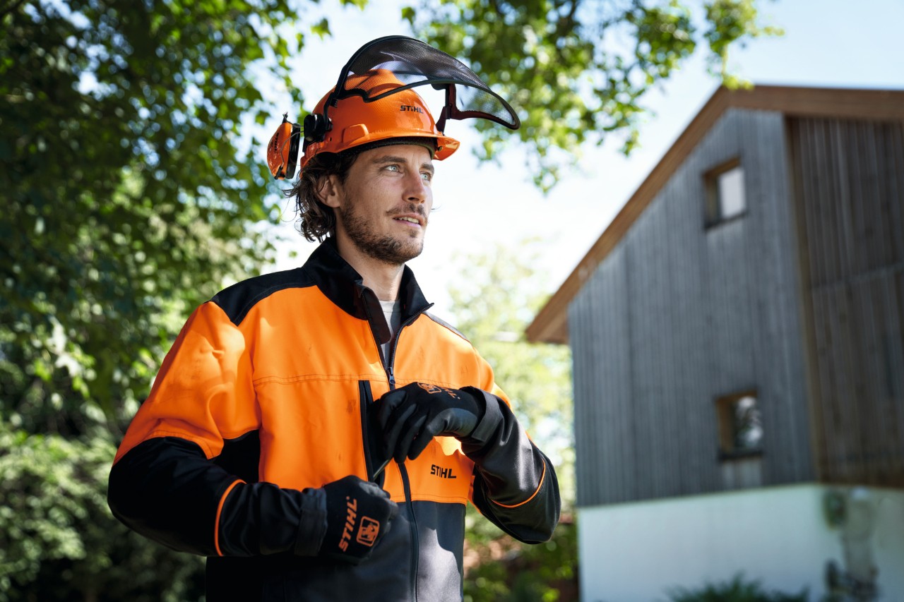 Un hombre con equipamiento de protección STIHL naranja y negro, incluido un casco y guantes, mira con confianza desde la imagen; en segundo plano, una casa y árboles 