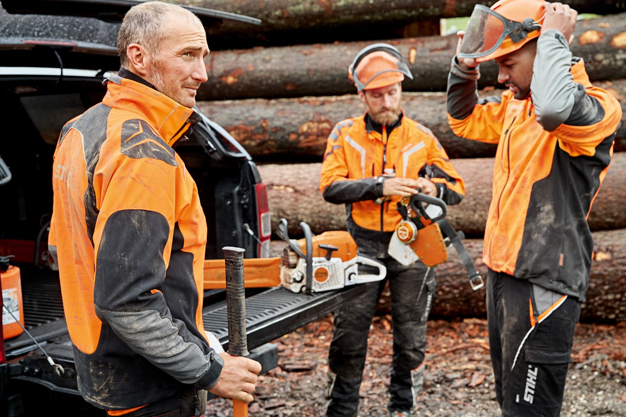 Tres hombres con ropa de protección STIHL delante de una pila de madera se preparan para trabajar con una motosierra STIHL