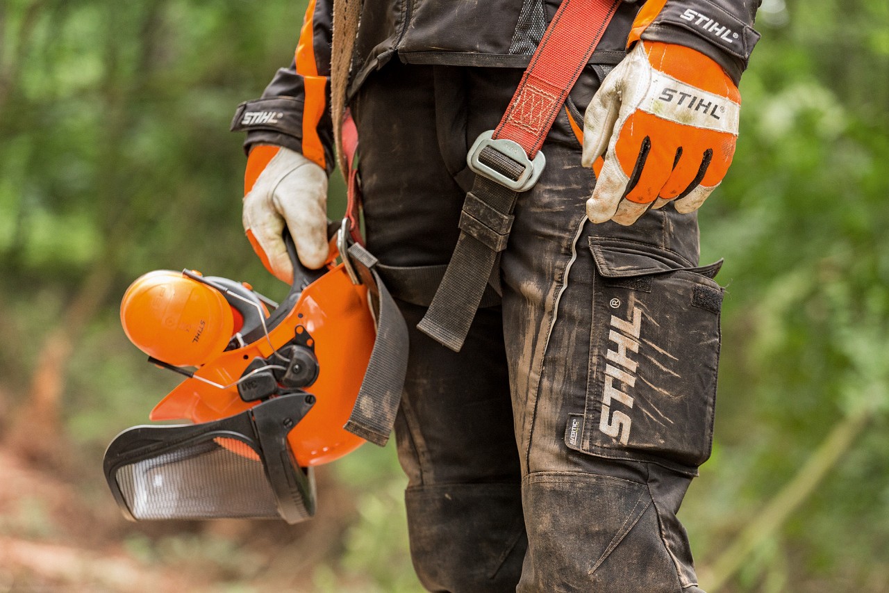 Una persona con protecciones para motosierras, lleva guantes y pantalones de trabajo STIHL sosteniendo un casco. 