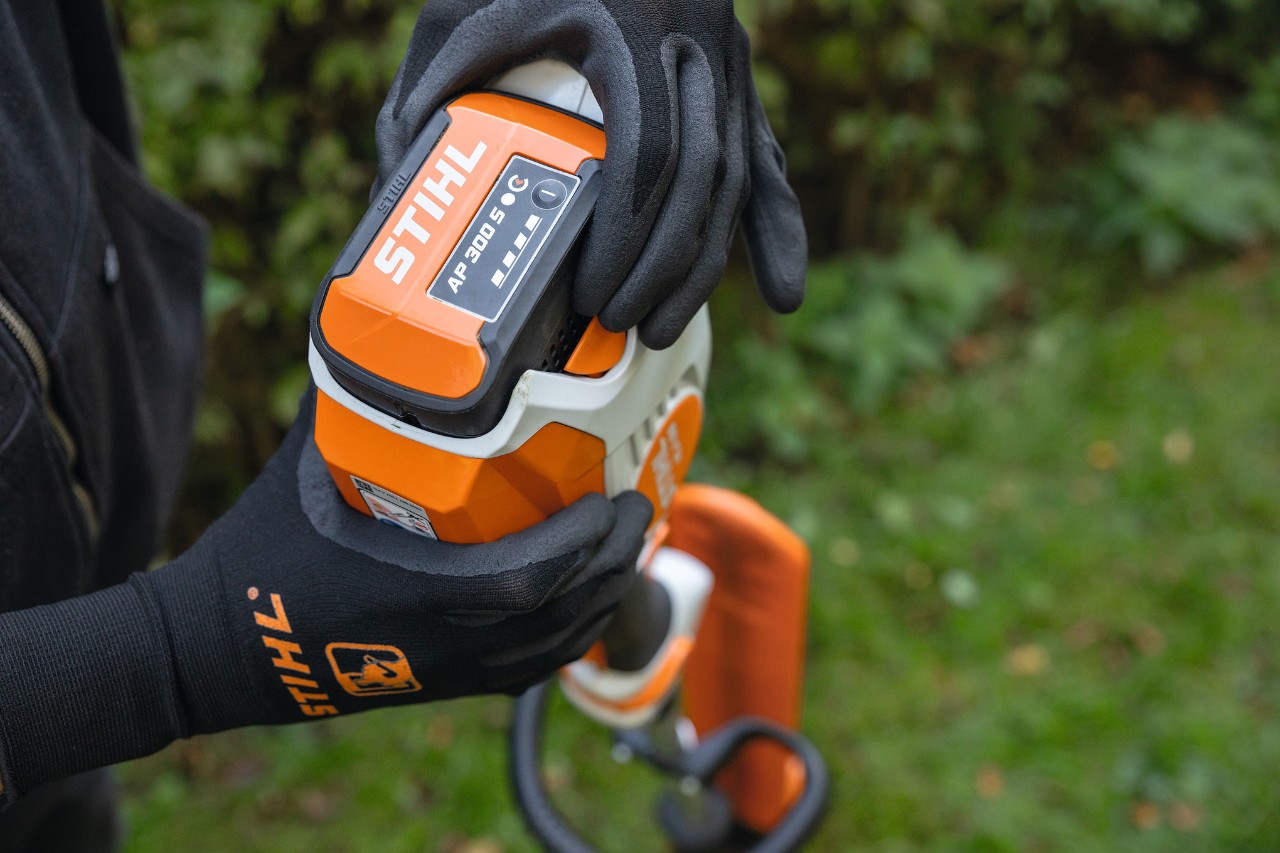 Primer plano de una persona con guantes de protección negros que agarra el cortasetos de altura a batería STIHL HLA 66 y trabaja con la batería de litio-ión STIHL AP 300 S
